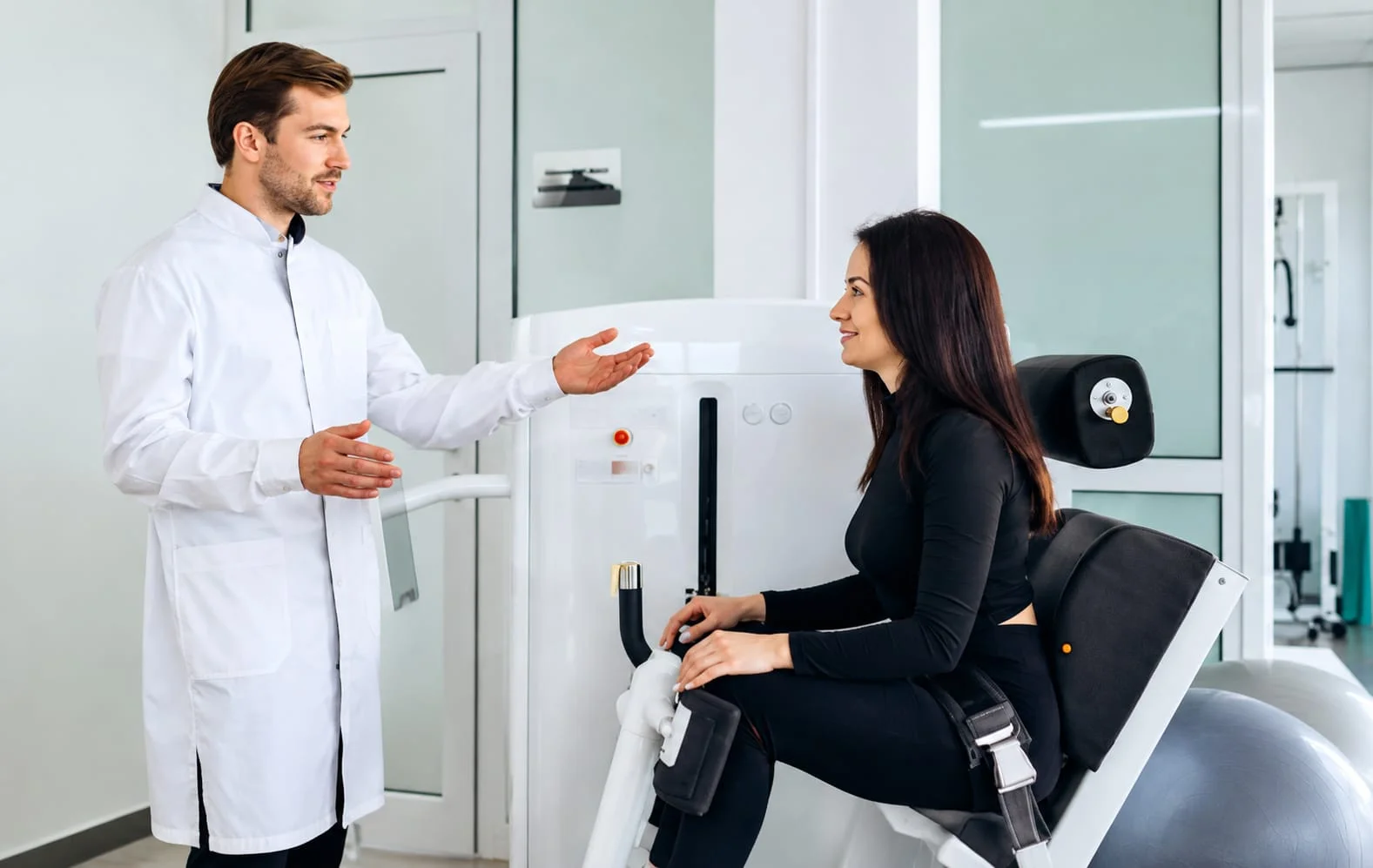 Physiotherapist talking with a woman seated on a rehabilitation exercise machine in a modern clinical setting.